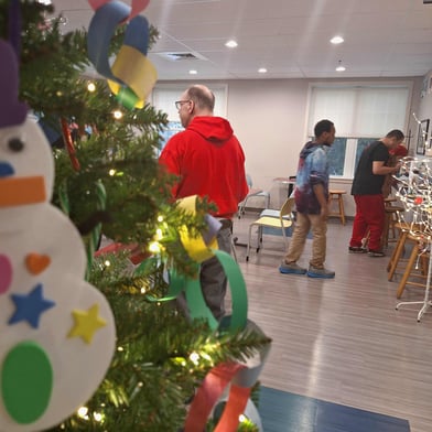 In the foreground is a Christmas tree lit up and decorated with handmade ornaments. In the background are students in the cafe decorating cookies.