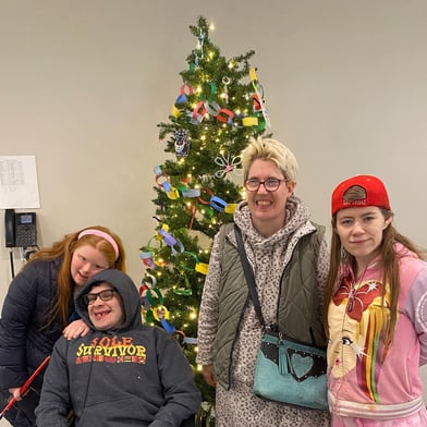 Four students stand in front of a Christmas tree in the cafe. On the left is a woman with red hair in a coat. Next to her is a man wearing a gray sweatshirt with hood on in a wheelchair. Next to him is a woman with short blonde hair wearing a vest over a gray sweater. On the right is a woman with long hair in a backeards red baseball cap and pink sweatshirt.
