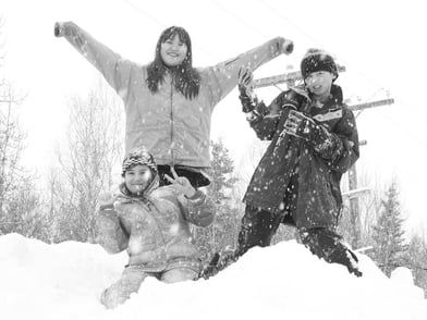 Black and white photo of three pre-teens playing in the snow while it's falling. The girl on the ground is giving a peace sign with her hand. The girl above her has her arms stretched wide. The boy on the right is playing air guitar.