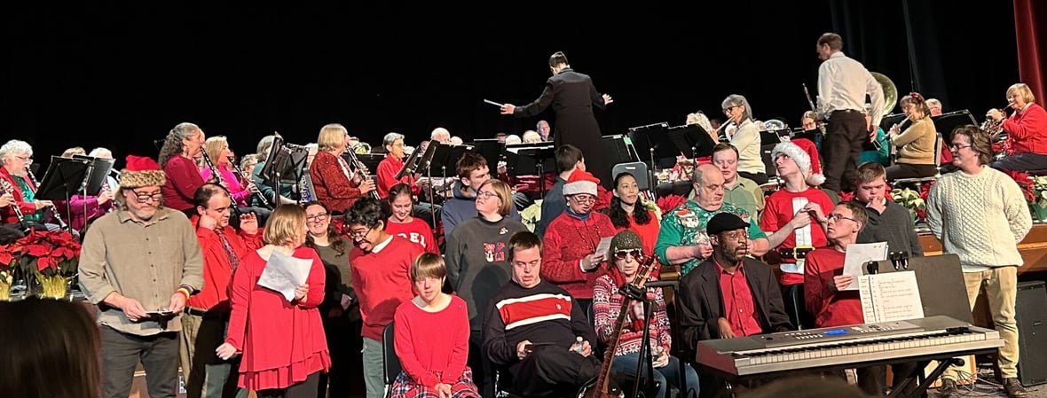 Joyful Chorus sits in front of the Florence Community Band on stage at Northampton High School for a winter show. The chorus is mostly wearing holiday sweaters. Whole Children and Milestones teachers Eddy (left) and Liam (right) stand on the ends of the group.